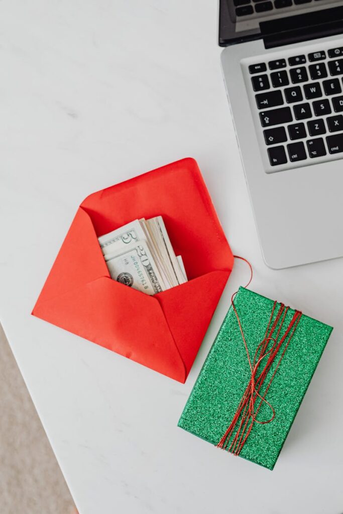 A red envelope containing cash beside a laptop and glittering green gift box on a white table.